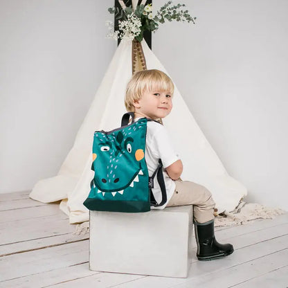 A boy sits on a green Dino-rrific kids backpack with a dinosaur face design. Ideal for outdoor adventures, featuring velvet and waterproof polyester materials, adjustable straps, and ample storage space.