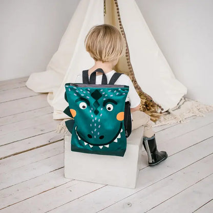 Child with backpack featuring Dino-rrific DINOSAUR design, sitting near green bag with face. Perfect for outdoor adventures, with velvet top, waterproof polyester bottom, and adjustable straps.