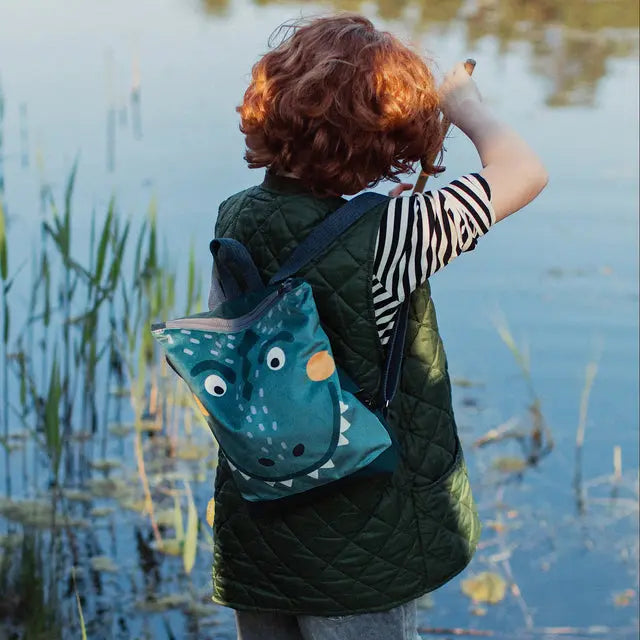 Child with Dino-rrific DINOSAUR kids backpack, gazing at fish in water. Velvet top, waterproof polyester bottom, adjustable straps, and ample storage for young adventurers.