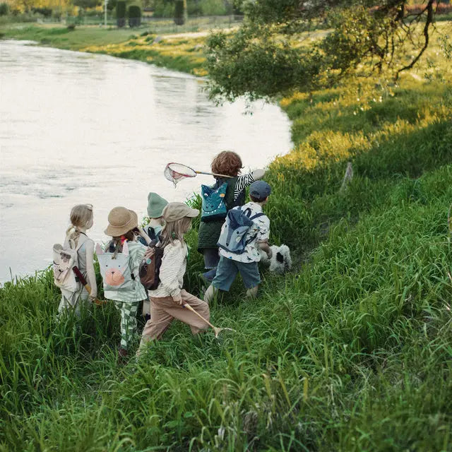 Children walking on grass by a river, one holding a stick, another wearing a Dino-rrific DINOSAUR kids backpack. Perfect for outdoor adventures, featuring a prehistoric design, adjustable straps, and ample storage space.