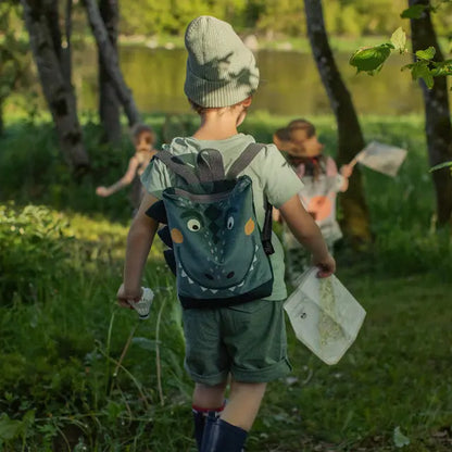 A child wearing a Dino-rrific DINOSAUR kids backpack, perfect for outdoor adventures. Features prehistoric design, velvet top, waterproof bottom, adjustable straps, and zip closure. Small: 29x26 cm, Large: 35x30 cm.