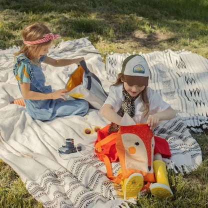 Two children with a girl opening a FOX backpack on a blanket outdoors. Velvet top with a Fox design, waterproof polyester bottom, adjustable straps, and zipper closure. Ideal for ages 3-9.