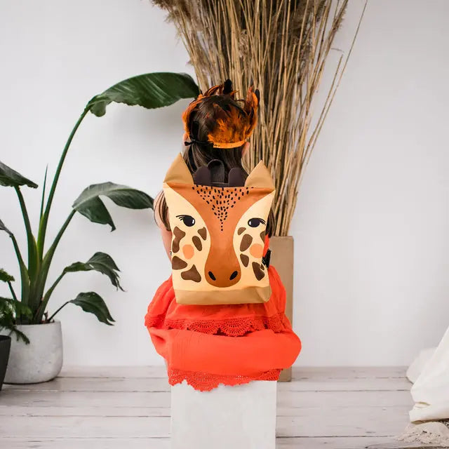 A child sits on a chair, holding a giraffe-themed backpack. The backpack features a velvet top with a giraffe design and sturdy polyester bottom in orange. Perfect for outdoor adventures!