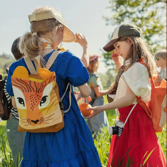 A group of children in a field, a girl with a tiger face backpack, and a girl holding an orange. Kids backpack - Giraffe, perfect for outdoor adventures.