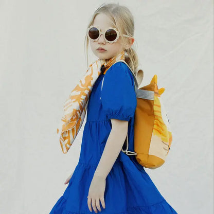 A girl in a blue dress and sunglasses, modeling a Kids backpack - Giraffe. Velvet top with giraffe design, waterproof polyester bottom, adjustable straps, and zipper closure.