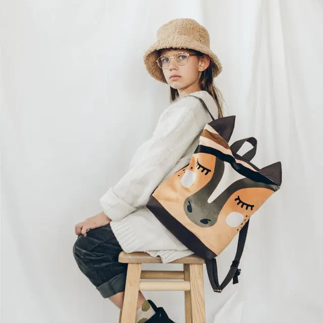 A girl sits on a stool with a backpack featuring a playful HORSE design. Lightweight and comfy, perfect for pre-school adventures. Measures 29cm x 26cm for ages 2-5.