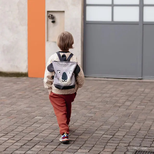 Child wearing a Koala backpack on a brick path, ready for outdoor adventures. Velvet top with Koala design, waterproof polyester bottom, adjustable straps. Small: H29cm x W26cm, Large: H35cm x W30cm.