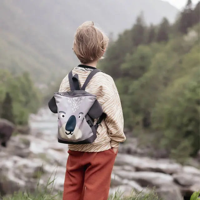 Child with koala-themed backpack by a river, ready for outdoor adventures. Velvet top, waterproof polyester bottom, adjustable straps, and secure zipper closure. Available in small and large sizes.