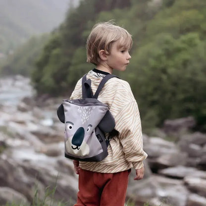 Child wearing a KOALA backpack outdoors, perfect for adventures. Velvet top with koala design, waterproof polyester bottom. Adjustable straps, zipper closure, ideal for 3-9-year-olds.