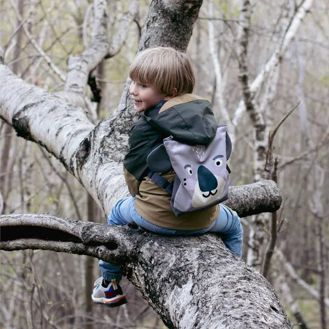 A child sits on a tree branch, holding a Kids backpack - Koala. The backpack features a cartoon face and velvet fabric with a koala design. Perfect for outdoor adventures with adjustable shoulder straps.