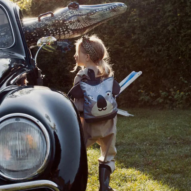 A child wearing a koala backpack, walking beside a car with a crocodile head. Perfect for outdoor adventures. Made of velvet and waterproof polyester. Adjustable straps for comfort.