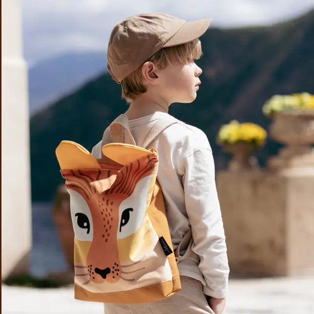 A boy wearing a lion-themed backpack with a hat, perfect for trips or playdates. Features a lion design, velvet top, waterproof polyester bottom, and adjustable shoulder straps.