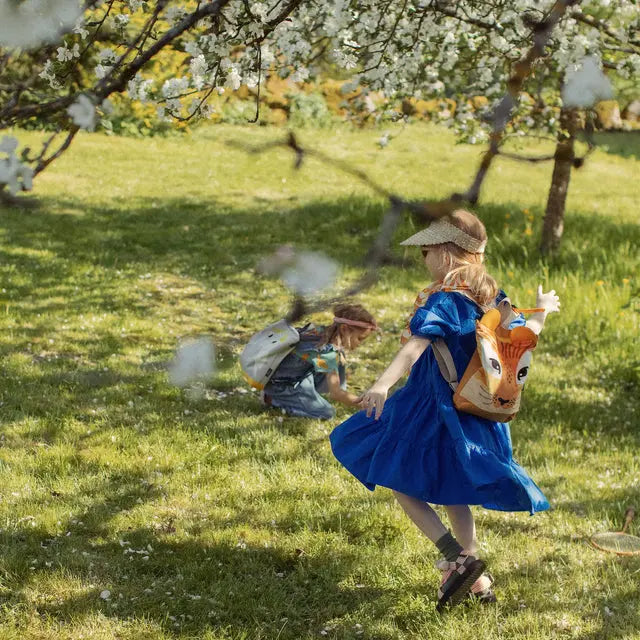 A young child in a blue dress and hat running on grass, carrying a lion-themed backpack. The backpack features a lion design, velvet top, waterproof polyester bottom, adjustable shoulder straps, and zipper closure.