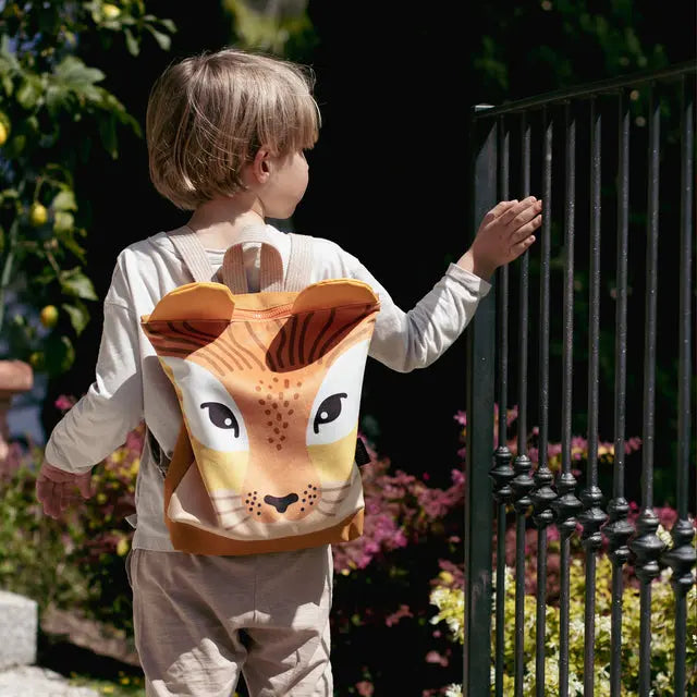 A child wearing a lion backpack with a face design, perfect for outdoor adventures. Made of velvet and waterproof polyester, featuring adjustable shoulder straps and zip closure. Two sizes available.