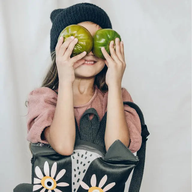 A girl playfully holds two tomatoes in front of her eyes, showcasing the Kids Backpack - Owl. The backpack features an adorable owl design, adjustable straps, velvet top, and waterproof polyester bottom. Perfect for young adventurers!