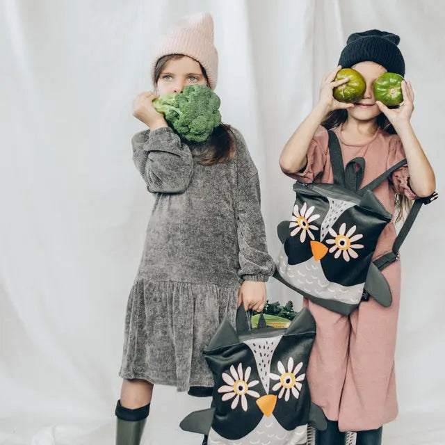 Two girls posing with vegetables, one in a grey dress and the other with a backpack. One holding a broccoli, the other a bag.