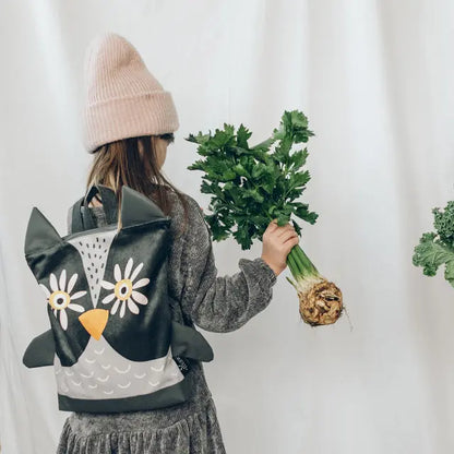 A girl holding a celery and a bag, wearing a hat and backpack, showcasing the Kids Backpack - Owl. Adjustable straps, velvet top, waterproof polyester bottom, zipper closure, perfect for preschool adventures.