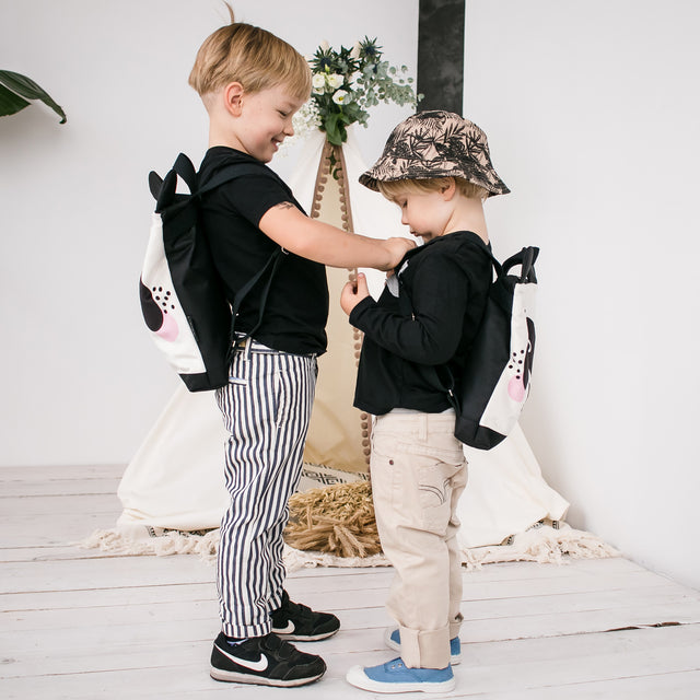 Kids backpack - Panda: Two boys stand by a teepee, one holding the panda backpack, showcasing its velvet top, polyester bottom, and adjustable straps. Ideal for young adventurers.