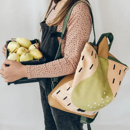 A woman carrying a pear-themed kids backpack with a velvet top and waterproof polyester bottom. Features adjustable beige shoulder straps, zipper closure, and two inside slip pockets. Ideal for young explorers aged 4-10.