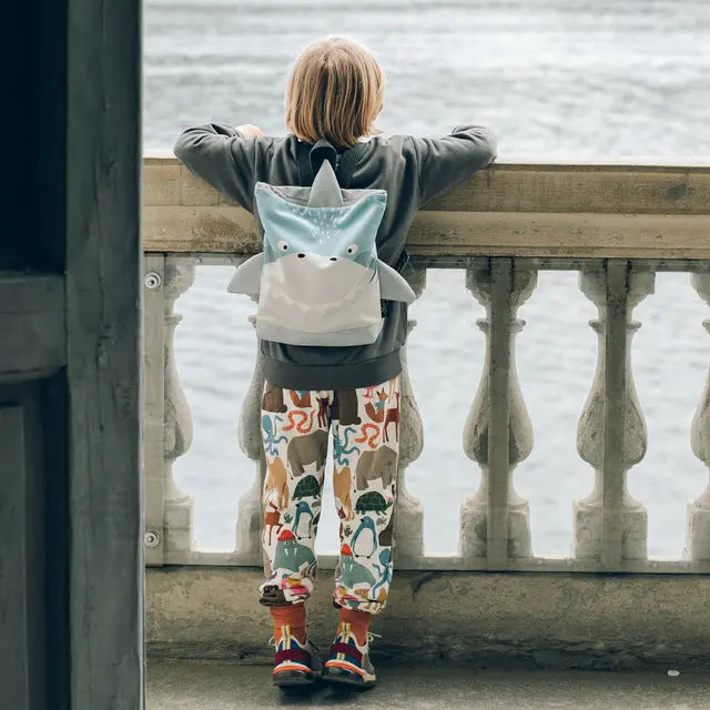 A child in pajamas with a shark backpack gazes at water. The SHARK backpack S/L for outdoor adventures, featuring a shark face design, velvet top, and waterproof polyester bottom.