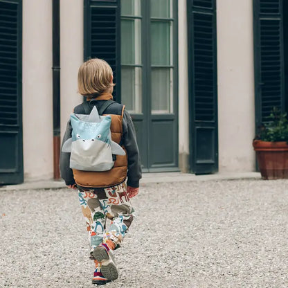 Child wearing a SHARK backpack in gravel area, with a plant in a pot in the background. Velvet top with shark design, waterproof polyester bottom, adjustable straps. Sizes: S (2-5 years), L (4-10 years).