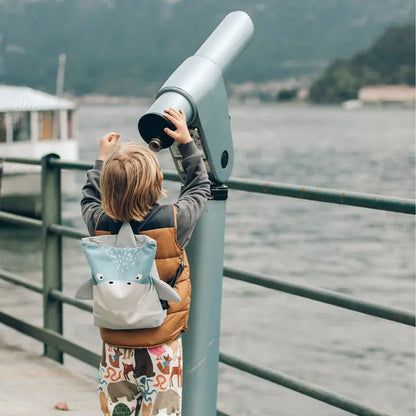 Child with shark backpack looking through telescope outdoors. Velvet top with shark design, waterproof polyester bottom. Adjustable straps, zipper closure. Small: 29x26cm, Large: 35x30cm.