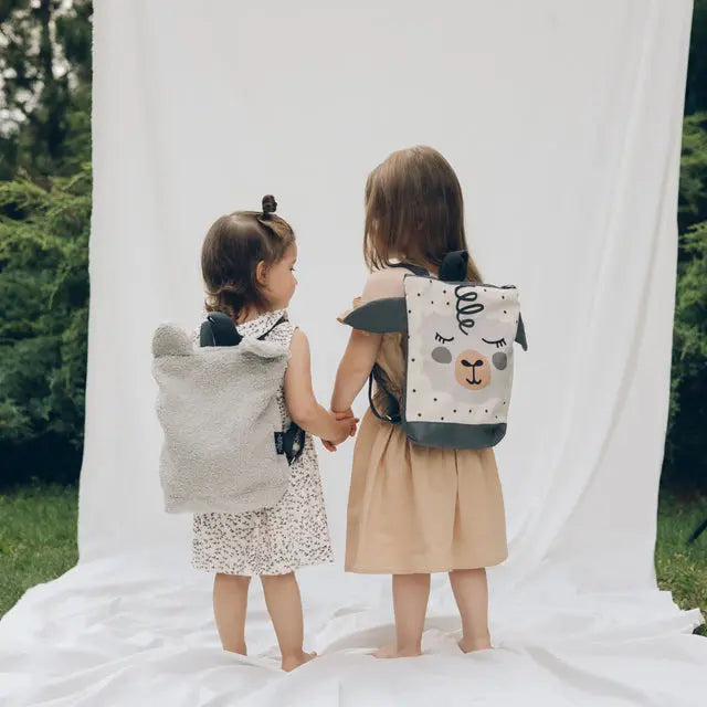 Two girls holding hands on a blanket, one wearing a white dress and a sheep-themed backpack. Kids backpack - Sheep, perfect for outdoor adventures with its fun design and ample storage.