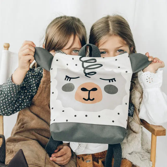 Two girls happily hold up a SHEEP backpack, featuring a playful sheep design. Ideal for young adventurers, with velvet and waterproof polyester materials, adjustable straps, and ample storage.