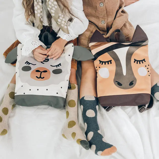 Two girls on a bed with bags, one sewing a llama pillow. A bag with a face on it. Kids backpack - Sheep, perfect for outdoor adventures.