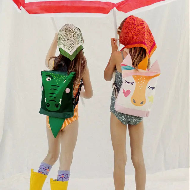 Two girls in swimsuits holding an umbrella, a child with a green backpack, and a unicorn-themed kids backpack with velvet top, waterproof bottom, and adjustable straps. Ideal for day-care, park, or hikes.