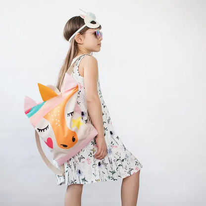 A girl in a white dress and sunglasses carries a unicorn backpack. Kids backpack - Unicorn: velvet top with unicorn design, waterproof bottom, adjustable straps, zipper closure, and pockets.