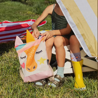 A child sits in a chair with a unicorn-themed backpack underneath. The backpack features a velvet top with a unicorn design and a waterproof pink bottom. Ideal for day-care, park outings, or hikes.