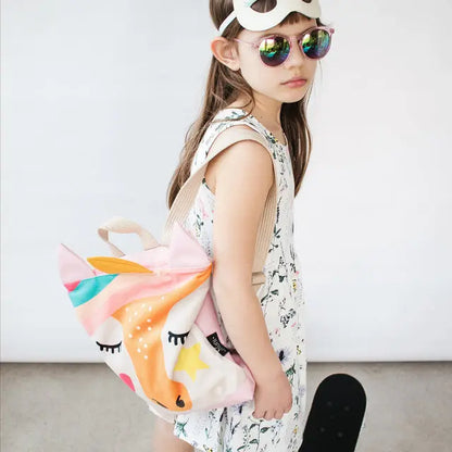 A girl in sunglasses and a mask holds a Kids backpack - Unicorn, featuring velvet top with Unicorn design, waterproof pink polyester bottom, adjustable straps, and zipper closure.