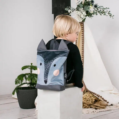 A child sits on a white cube with a backpack featuring a fun wolf design. The backpack is made of velvet and waterproof polyester, with adjustable straps and inside pockets. Height: 29-35cm.