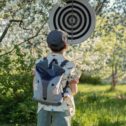 A child standing in a field with a dart board, holding the Kids backpack - Wolf. Multifunctional velvet and polyester backpack with wolf design, perfect for young explorers.