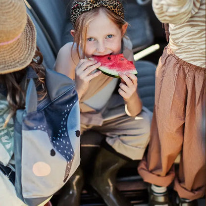 A girl in a sun hat eating watermelon, showcasing the Kids backpack - Wolf. Multifunctional velvet and polyester backpack with a fun wolf design for young explorers.