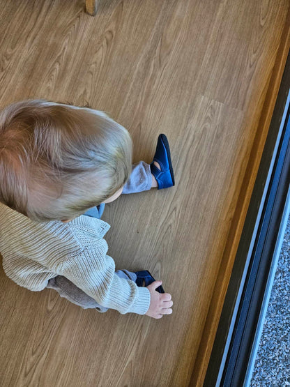 Child wearing Kids' Slippers Mutsu - Blue, featuring a wide toe box, air holes, and adjustable Velcro, sitting indoors, showcasing the slipper's soft leather upper.