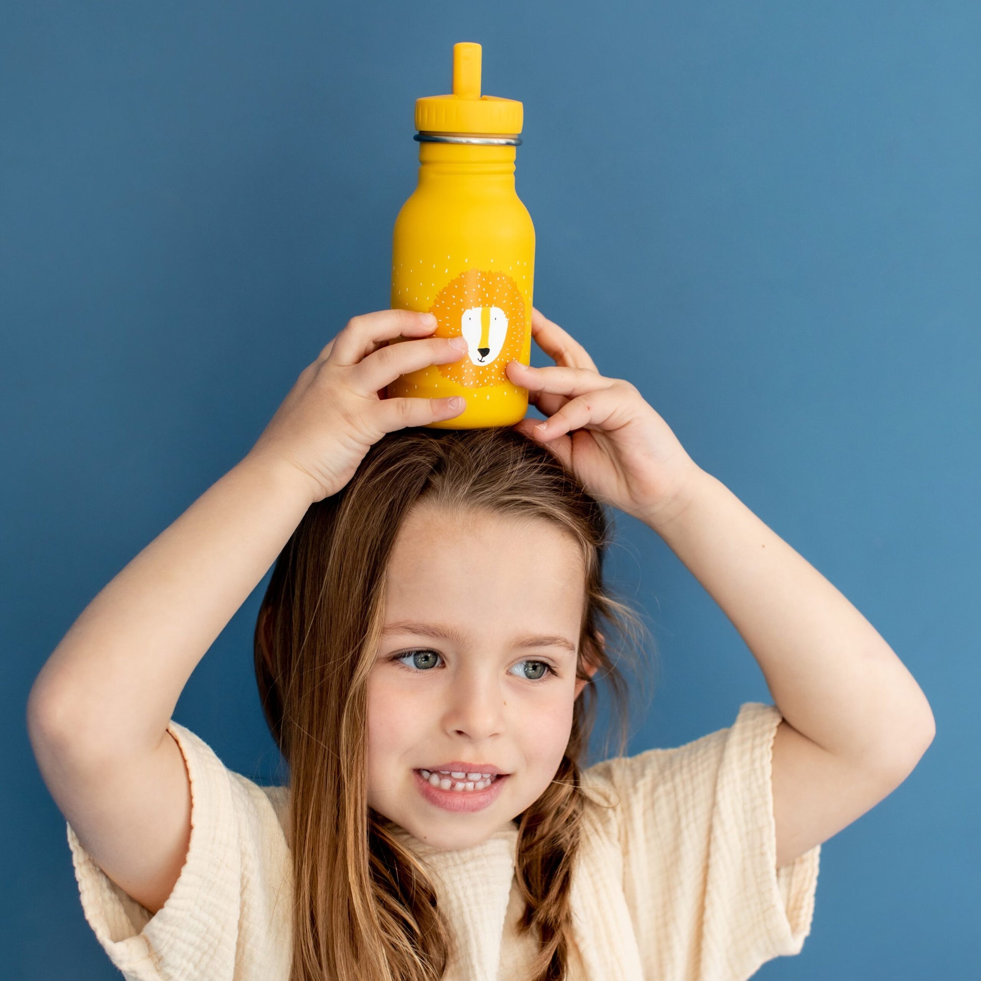 A girl holds a Stainless Steel Bottle 350 ml - Mr Lion on her head. Durable, leak-proof, stainless steel bottle with kid-friendly cap and loop for easy carrying. Dimensions: 15.5 cm x 6.5 cm.