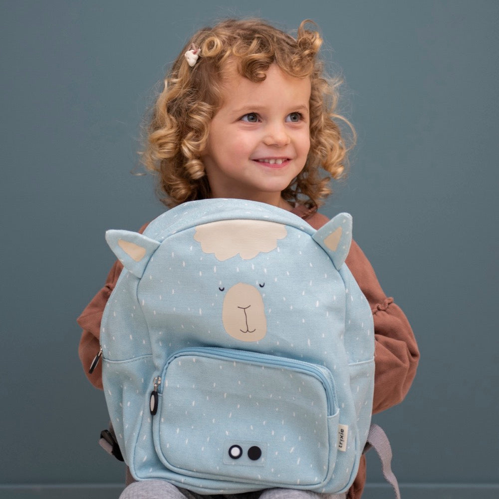 Child with curly hair holding Mr Alpaca backpack, featuring adjustable straps, chest strap, water repellent cotton material, and 7.5L capacity for ages 3+. Dimensions: 31 x 23 x 10 cm.