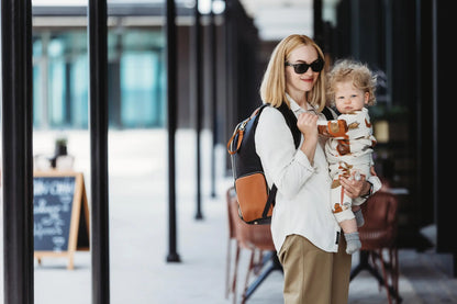 A woman wearing sunglasses holds a baby while showcasing the Large Diaper Backpack – Black Coffee. Designed for convenience with 16 pockets, laptop and baby food compartments, stroller straps, and a waterproof changing mat.