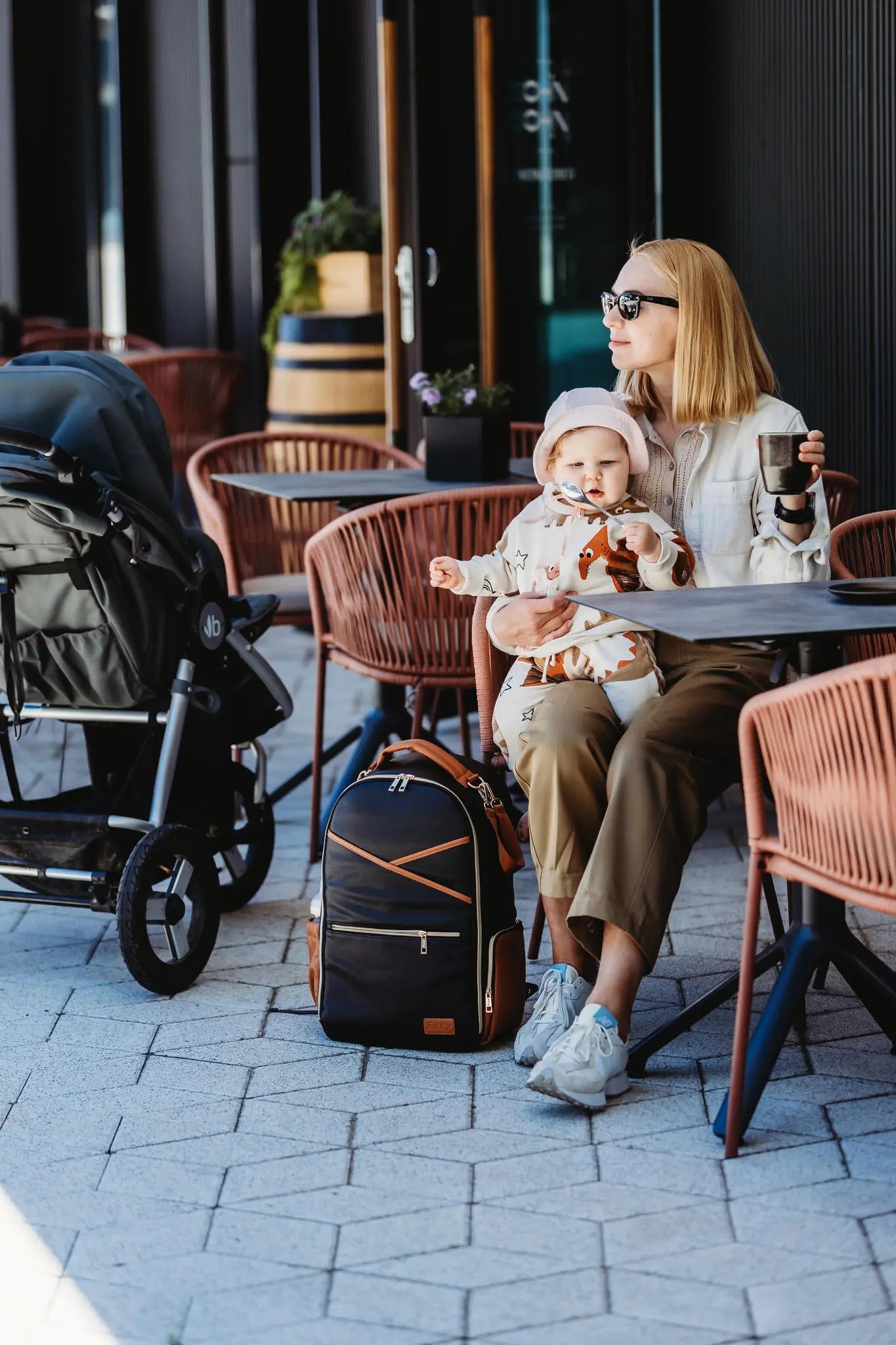 A woman sits with a baby next to a black and brown Large Diaper Backpack – Black Coffee. The backpack features 16 pockets, stroller straps, and a laptop pocket for ultimate convenience on the go.