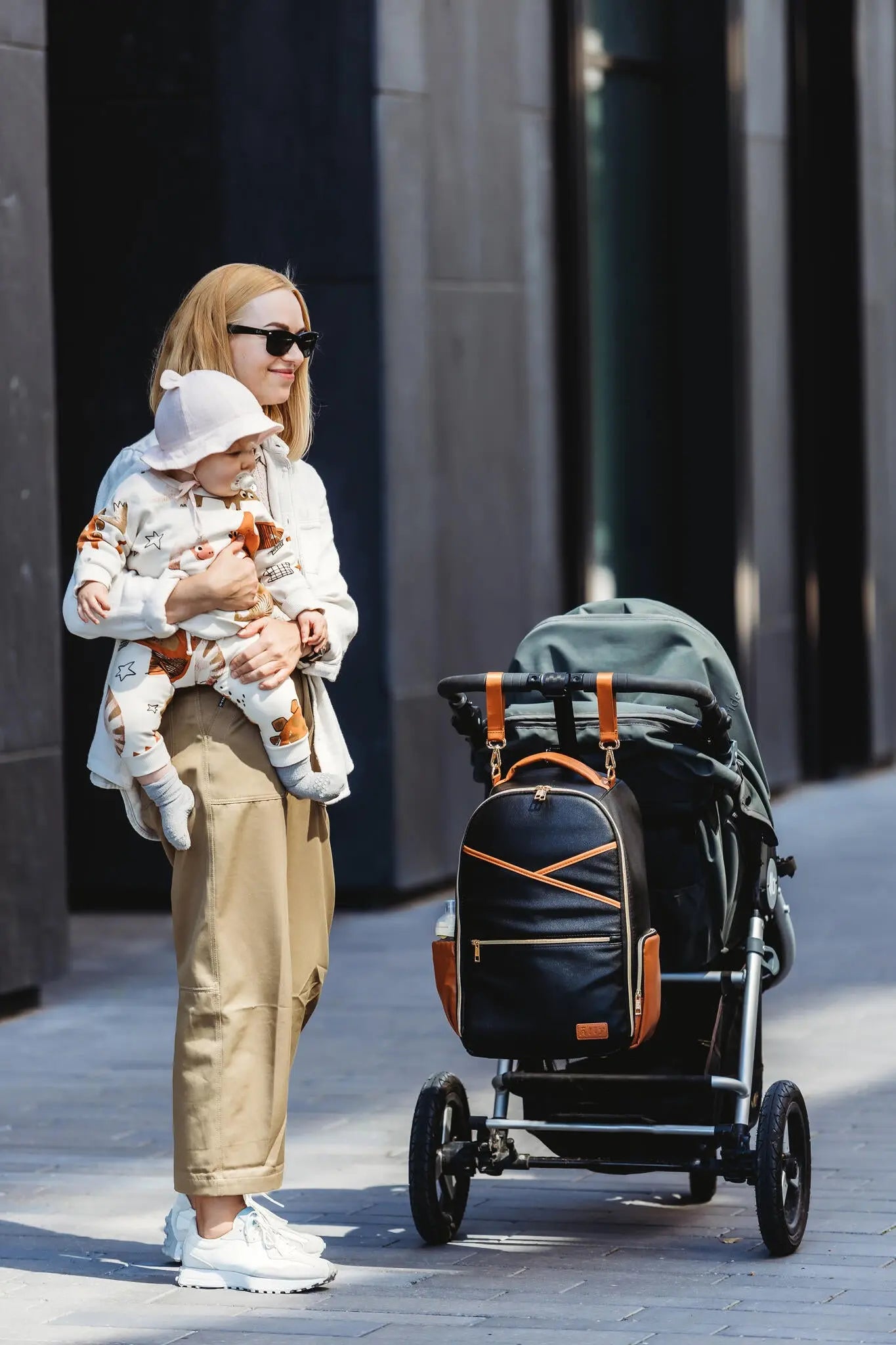 A woman holds a baby next to a stroller with a backpack attached. A black coffee diaper bag, featuring 16 pockets, stroller straps, laptop and baby food compartments, and a waterproof changing mat.