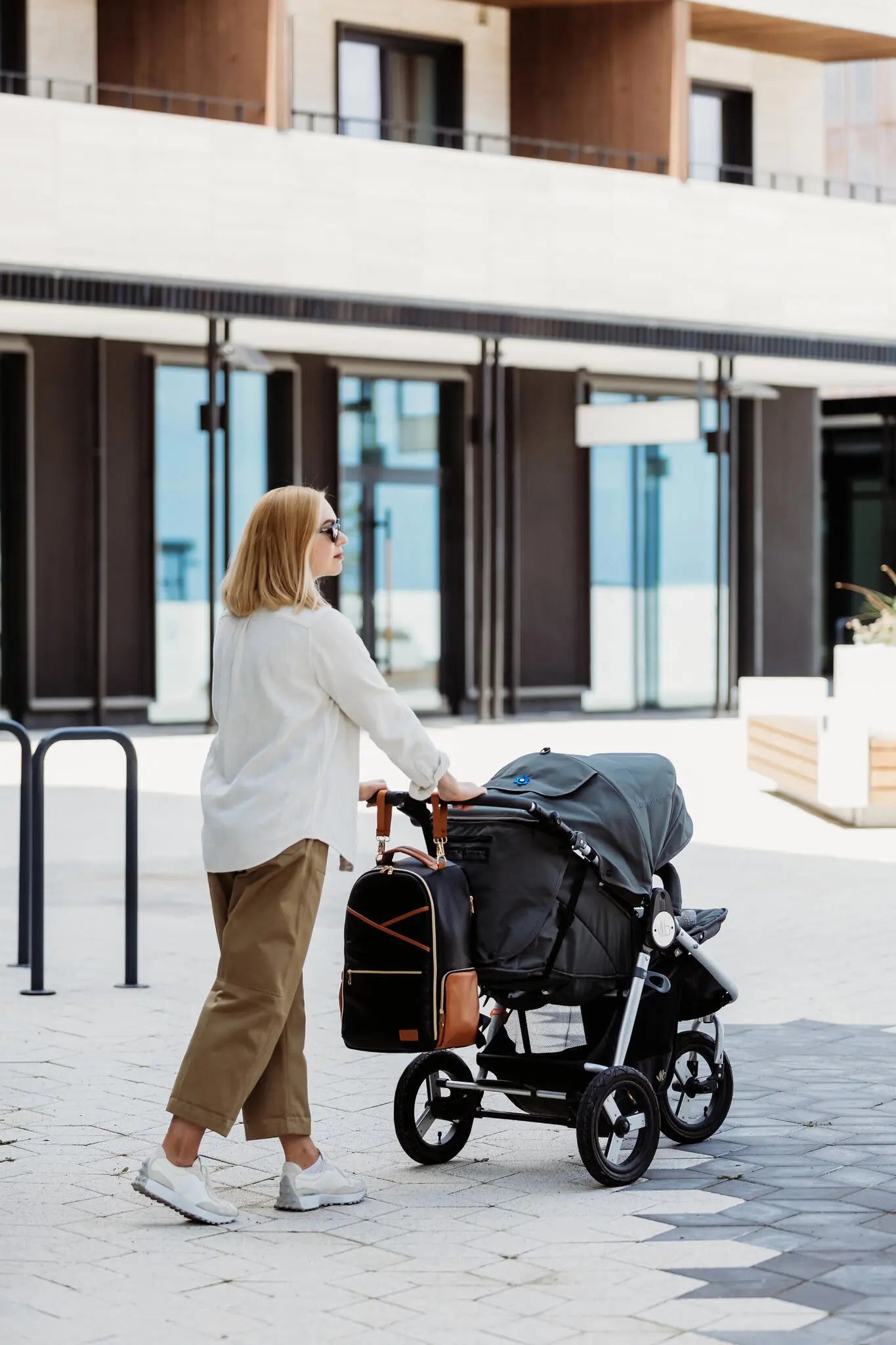 A woman with blonde hair pushing a stroller, showcasing a spacious Black Coffee Diaper Bag with 16 pockets, laptop and baby food compartments, stroller straps, and waterproof changing mat.