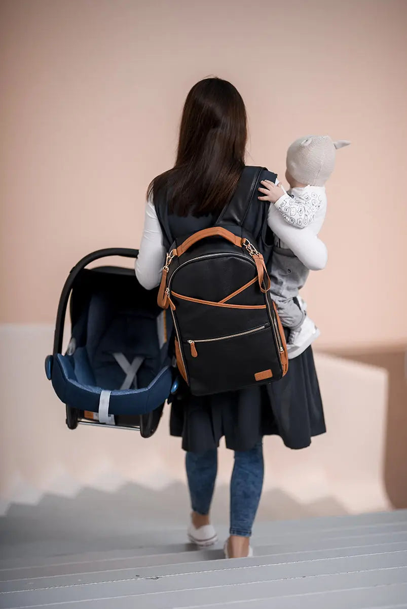 A woman carrying a baby in a spacious Black Coffee Diaper Bag with 16 pockets, laptop and baby food compartments, stroller straps, and a waterproof changing mat.