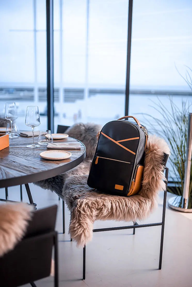 A spacious Black Coffee Diaper Backpack on a chair, showcasing a stylish and organized design for parents on the go. Features 16 pockets, laptop and baby food compartments, stroller straps, and lightweight vegan leather.