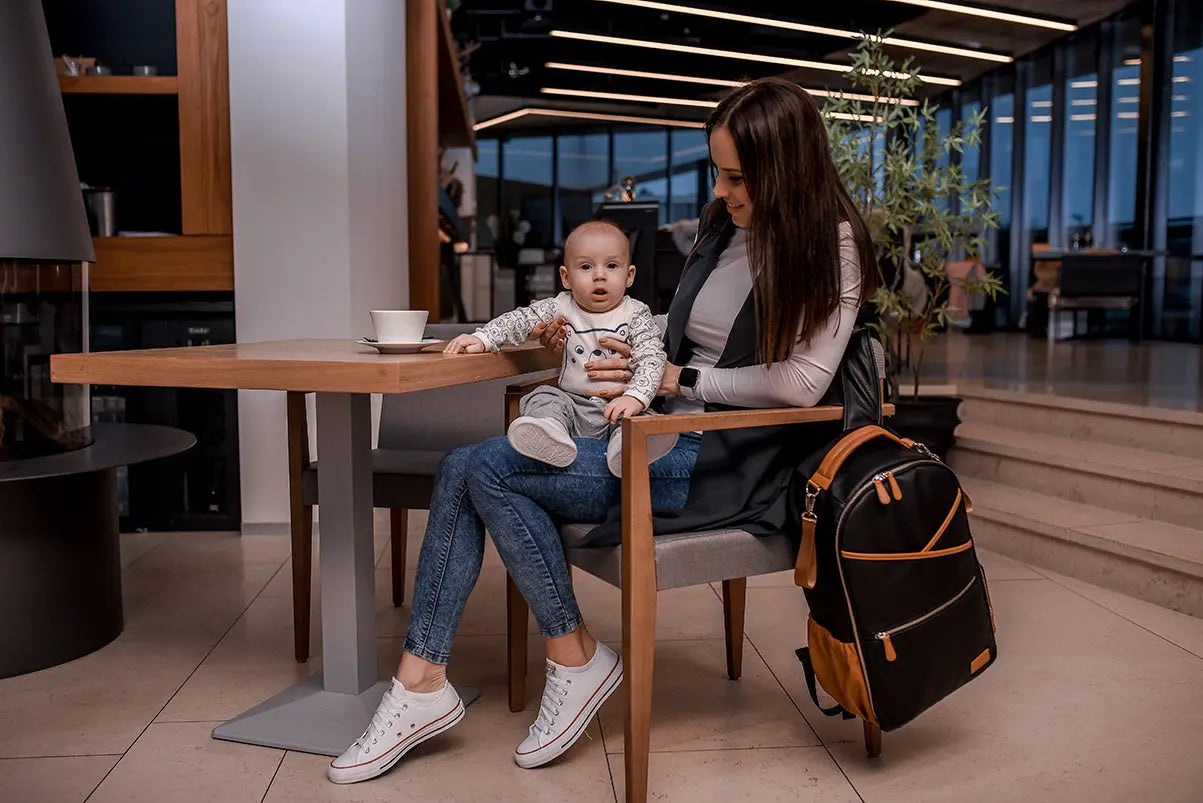 A woman sits holding a baby, showcasing the Large Diaper Backpack – Black Coffee. Designed in Europe, it features 16 pockets, stroller straps, and a laptop pocket for ultimate convenience.