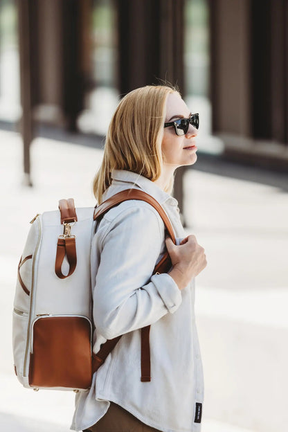 A woman wearing sunglasses carries a spacious Cappuccino Diaper Bag with 16 pockets, laptop and baby food compartments, stroller straps, and a waterproof changing mat.