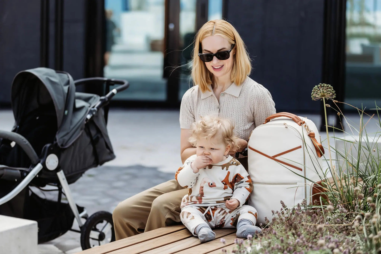 A woman sits on a bench with a baby, showcasing the Cappuccino Diaper Bag. Stylish, spacious design with 16 pockets, laptop and baby food compartments, stroller straps for hands-free convenience.