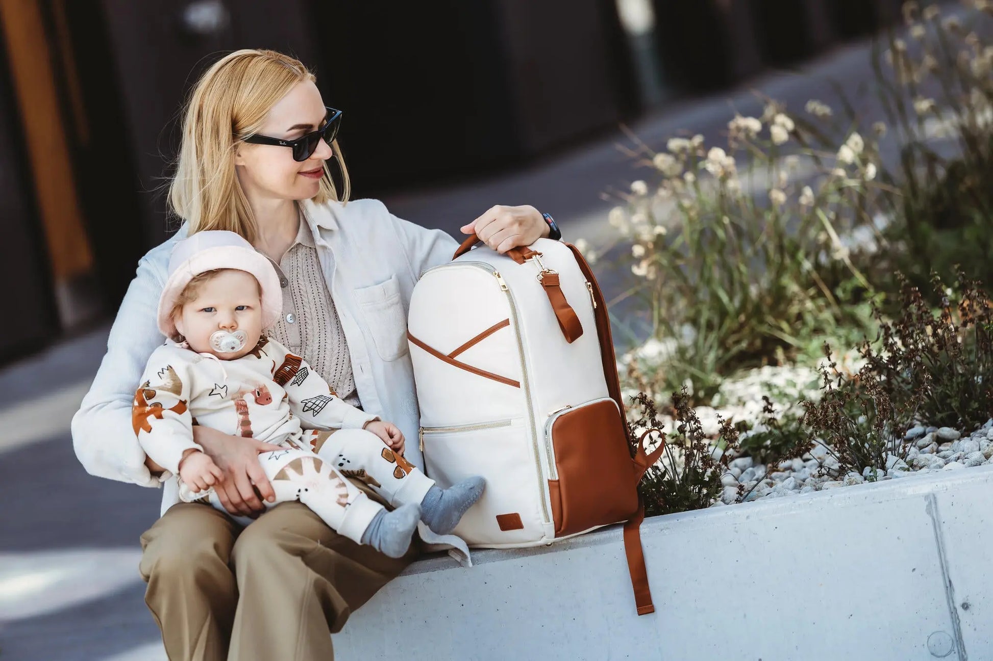 A woman holds a baby while carrying a spacious Cappuccino Diaper Bag with 16 pockets, laptop and baby food compartments, stroller straps, and a waterproof changing mat.