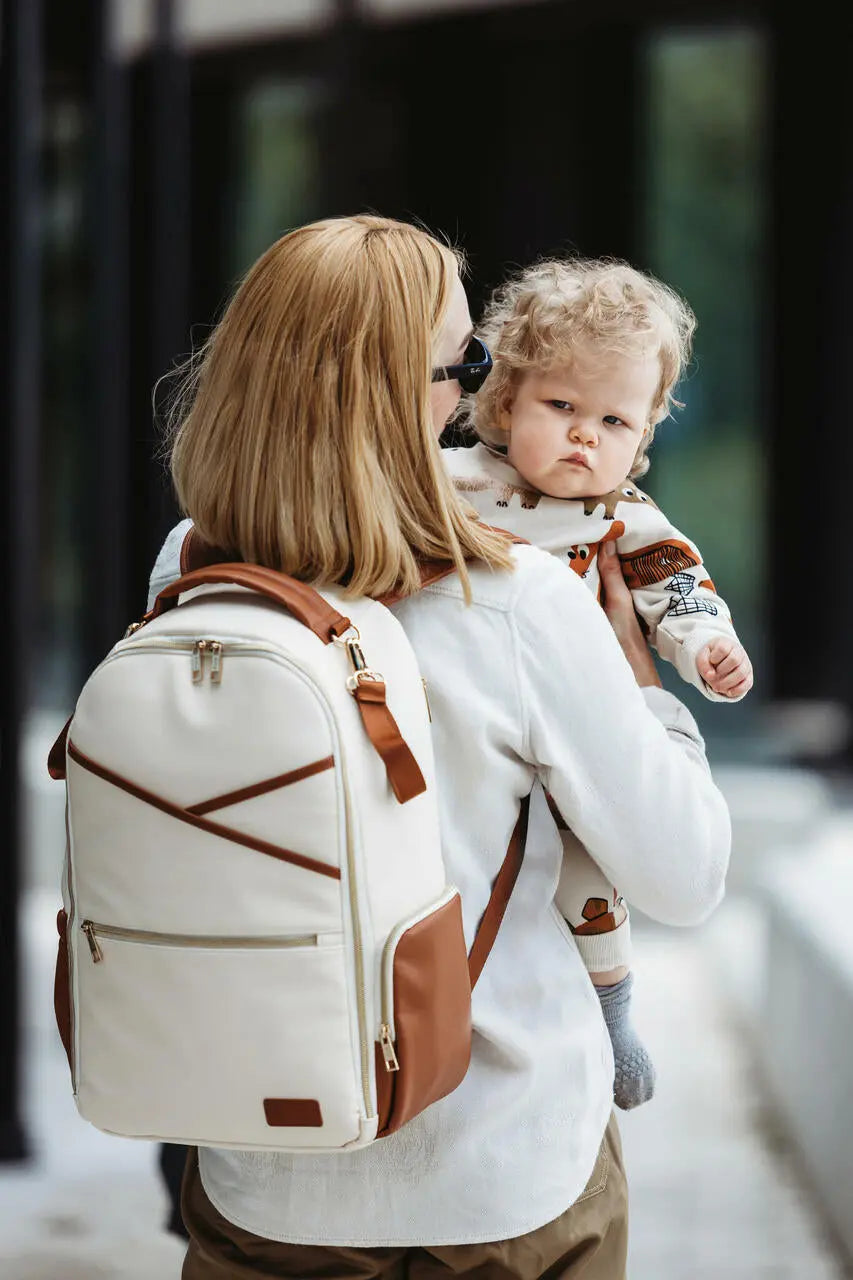 A woman carries a baby while showcasing the spacious Cappuccino Diaper Bag by Ally Scandic. Designed for style and functionality, featuring 16 pockets, stroller straps, and eco-friendly materials.